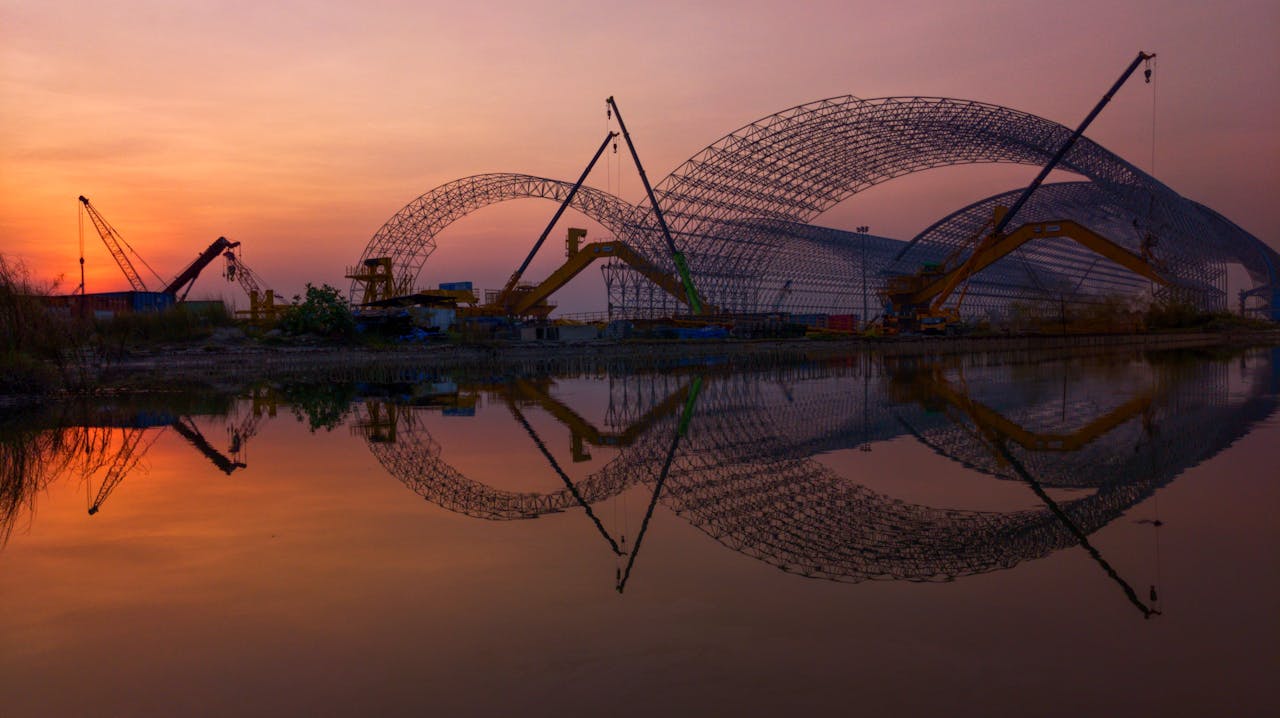 Silhouetted cranes at sunset reflect in water, capturing construction in Khulna, Bangladesh.